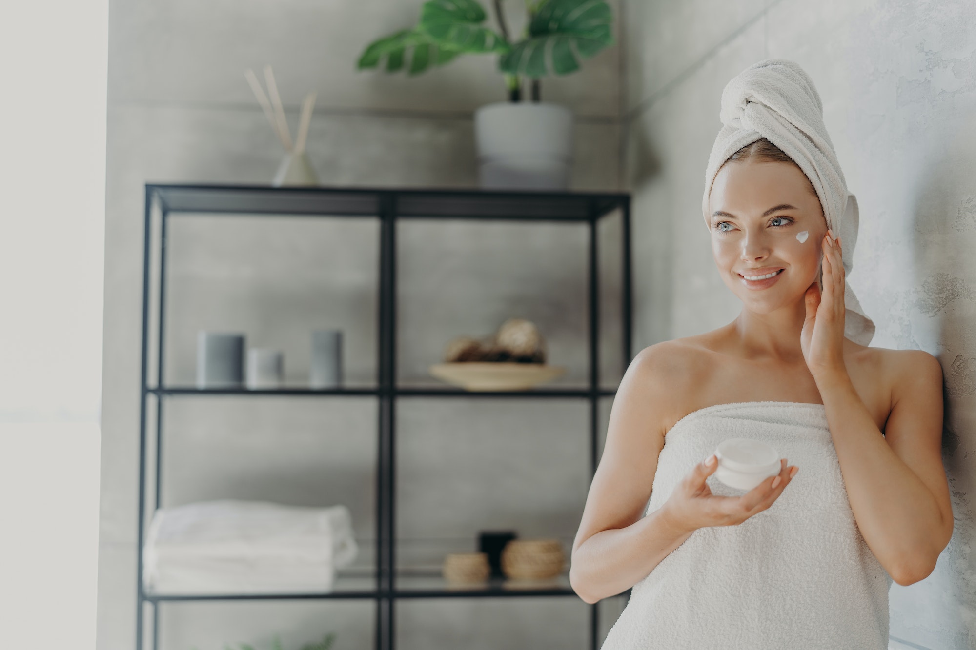 Lovely smiling Caucasian woman applies beauty cream on cheek, enjoys morning domestic skin care